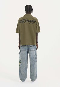 Olivgrön kortärmad skjorta med broderad "Ed Hardy" logotyp på baksidan, ihopparad med ljusblå baggy jeans med ett färgglatt mönster.