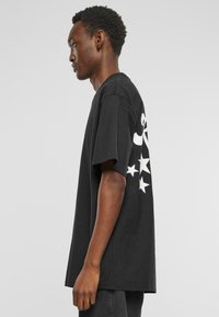 Black cotton t-shirt with oversized fit, featuring white graphics and stars on the left sleeve. Rounded neckline and short sleeves.