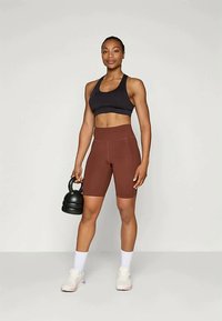 Αθλητική γυναίκα που στέκεται κρατώντας μια μαύρη kettlebell, φορώντας μαύρο αθλητικό σουτιέν, καφέ σορτσάκια ποδηλάτου με ψηλή μέση, λευκές κάλτσες και λευκά αθλητικά παπούτσια.