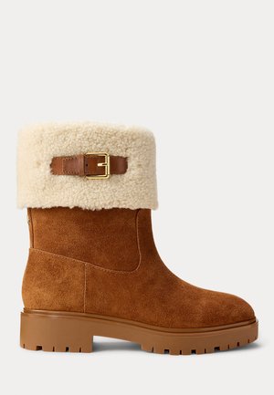 Brown suede ankle boot with a cream shearling cuff and a brown buckle accent. Features a chunky rubber sole and side zipper closure.