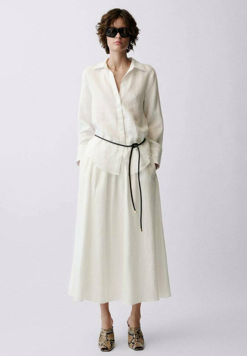 Ensemble chemise et jupe en lin blanc, avec une ceinture noire. Accessoirisé avec des lunettes de soleil noires et des chaussures à imprimé animal. Texture lisse, design fluide.