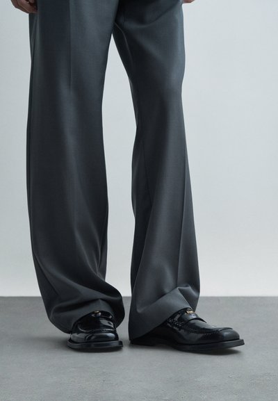 Eine Person trägt weit geschnittene graue Hosen und polierte schwarze Leder-Loafer. Sie steht auf einem grauen Boden vor einem schlichten Hintergrund.