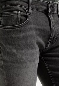 Zbliżenie na ciemnoszare dżinsy pokazujące przednią kieszeń, pętlę na pasek, guzik i zamek błyskawiczny.