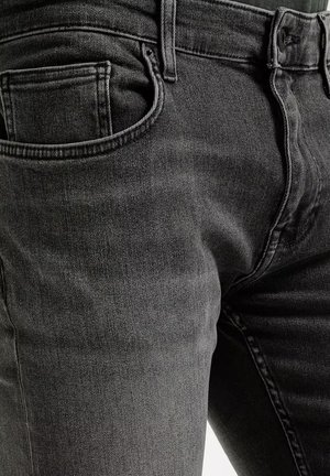 Zbliżenie na ciemnoszare dżinsy pokazujące przednią kieszeń, pętlę na pasek, guzik i zamek błyskawiczny.