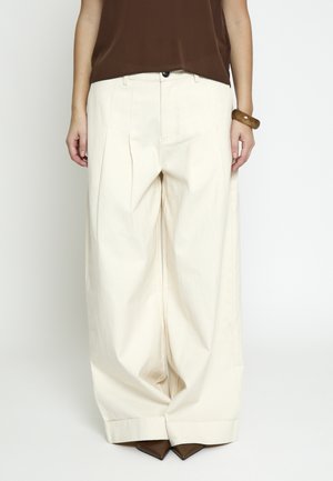 Kobieta ubrana w kremowe spodnie o szerokich nogawkach, brązowe szpiczaste buty, brązową bluzkę i brązową bransoletkę, stojąca na białym tle.