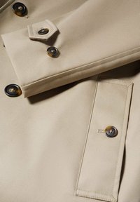 Manche et détail avant d'un trench-coat beige avec des boutons en écaille foncée et un rabat de poche cousu sur un tissu lisse.