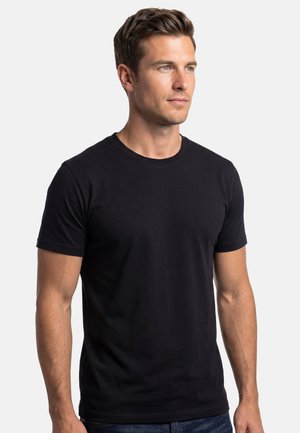 Młody mężczyzna z krótkimi brązowymi włosami, ubrany w prosty czarny t-shirt z krótkim rękawem, patrzący lekko w prawo na białym tle.