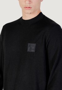 Maglione nero a maglia con scollo rotondo, caratterizzato da una toppa in pelle testurizzata sul petto con dettagli del logo. Tessuto morbido e testurizzato.