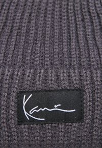 Grau gestrickte Mütze mit vertikalen Rippen. Ein schwarzes Etikett mit weißem Signaturdetail ist seitlich angenäht. Weiche Textur.