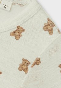 Ljusbeige tyg med en mjuk textur, prydd med ett upprepande mönster av små bruna nallebjörnar och en etikett vid halslinningen.