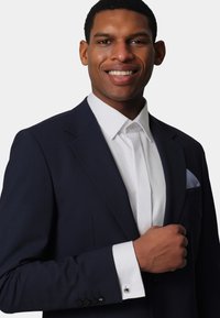 Dark navy suit with a notched lapel, paired with a white shirt. Cufflinks accentuate the white cuff. Pocket square features a blue check pattern.