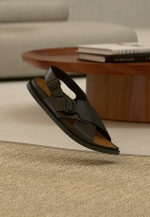 Schwarze Ledersandale mit zwei verstellbaren Schnallen, offenem Zehendesign und braunem Fußbett, dargestellt über einem strukturierten Teppichboden.