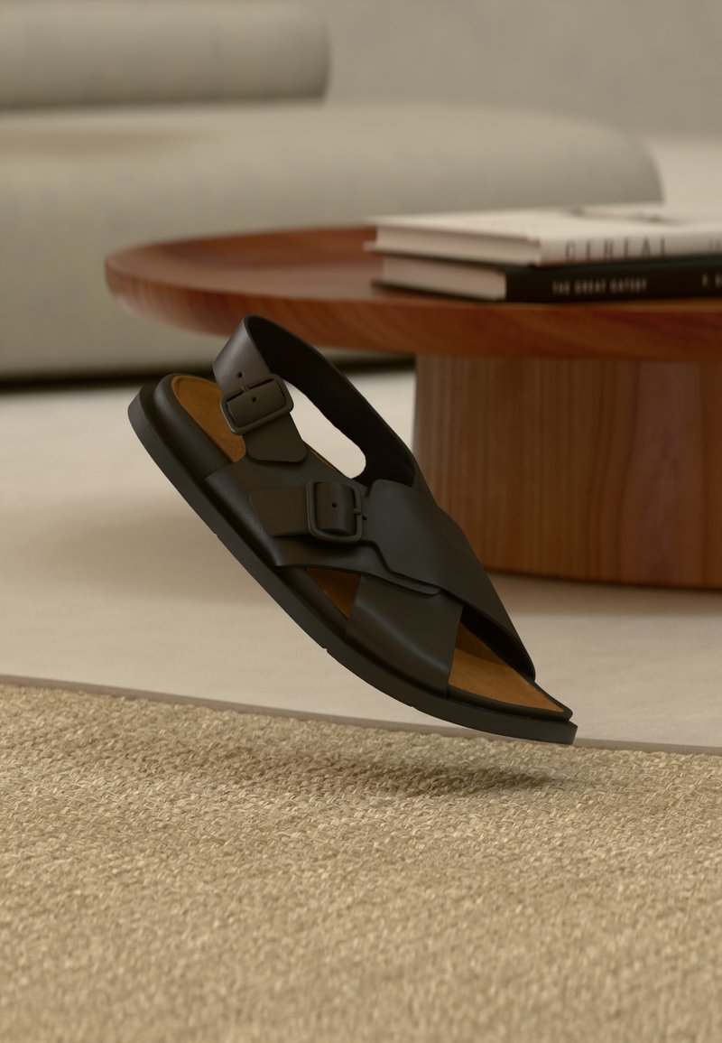 Sandale en cuir noir avec deux boucles réglables, design à bout ouvert et une semelle intérieure marron, présentée au-dessus d'un tapis texturé.