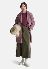 Kleid mit Fischgrätenmuster in Burgunderrot, mit Revers, olivgrüne Rock, lila Rollkragenpullover und cremefarbene Sneakers. Hält ein flauschiges, helles Accessoire.
