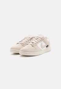 Par de ténis Nike de beige claro com laterais em malha branca, design com atacadores e etiqueta de tecido "Cordura" preta em fundo branco limpo.