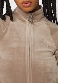 Beige zip-up velourjacka med hög krage, med en silverfärgad logotyp och dragkedja, som visar en mjuk textur och en subtil lyster.