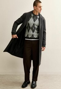 Man met een donkergrijze lange jas, een grijze argyle trui, donkerbruine broek en zwarte schoenen, met de jas openhoudend en naar de zijkant kijkend.