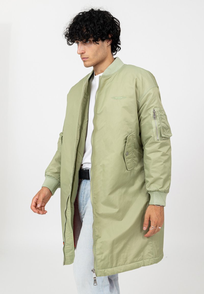 Jonge man met krullend zwart haar, gekleed in een lichtgroene lange bomberjack, een wit shirt, lichtblauwe jeans en een zwarte riem, staand tegen een witte achtergrond.