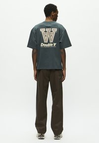 Übergroßes dunkelgrünes Baumwoll-T-Shirt mit einem großen cremefarbenen Grafikdesign und dem Text "W" sowie "Double A" auf der Rückseite, kombiniert mit braunen Hosen.