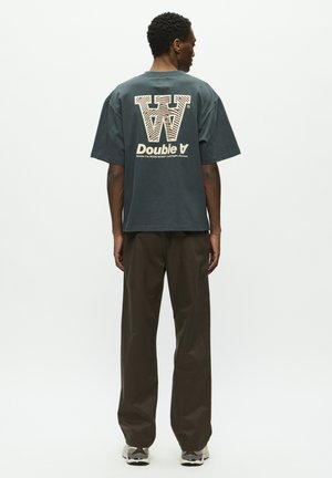 Oversized dark green cotton t-shirt featuring a large cream graphic with "W" and "Double A" text on the back, paired with brown pants.
