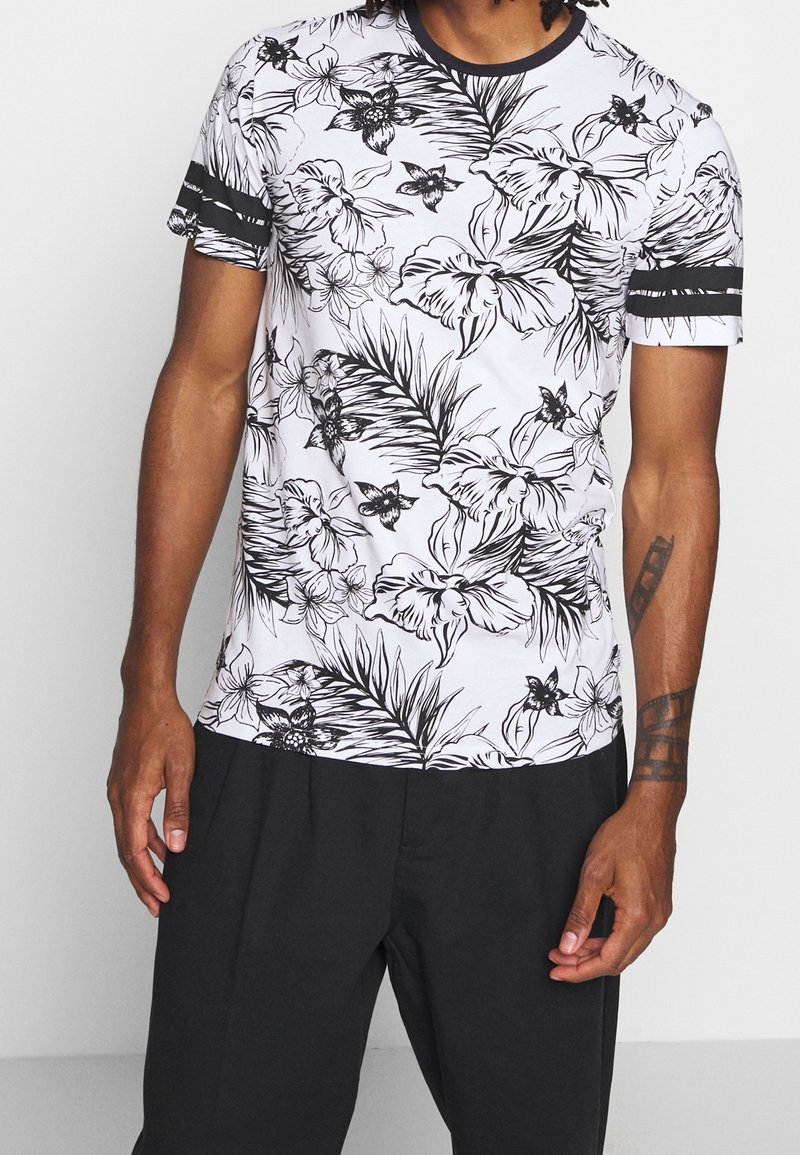 Homme portant une chemise blanche à manches courtes avec des motifs floraux et feuillages noirs et deux bandes noires sur chaque manche, assortie à un pantalon noir.