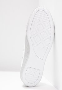 Semelle de sneaker blanche présentant un motif de bande de roulement texturé avec des formes géométriques, appuyée contre une surface d'angle blanche.
