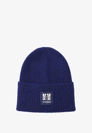Strikket hue i mørkeblå med en ribbet tekstur; har en rektangulær etiket med "STUDIO" og et dekorativt logo.