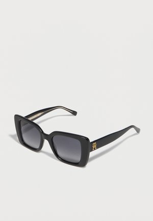 Store sorte firkantede solbriller med mørke tonede glas og guldlogo på stangen, vist på en lys baggrund.