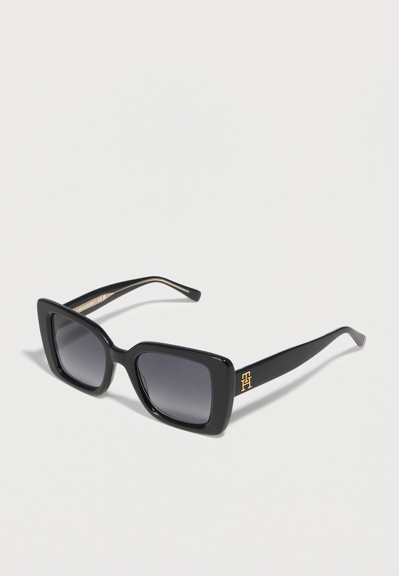 Μαύρα υπερμεγέθη τετράγωνα γυαλιά ηλίου με σκοτεινούς φακούς και χρυσό λογότυπο στο μπράτσο, σε ανοιχτό φόντο.