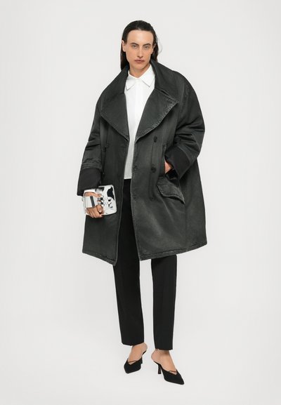 Άτομο που φοράει υπερμεγέθη σκούρο γκρι παλτό πάνω από λευκό πουκάμισο και μαύρο παντελόνι, κρατώντας μια μικρή ασημένια τσάντα, στέκεται μπροστά από μια απλή επιφάνεια.