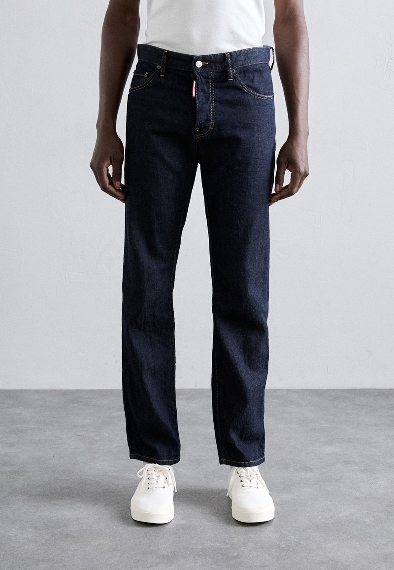Dunkelblaue Jeans mit geradem Schnitt, ausgestattet mit fünf Taschen, gelben Nähten und einem silbernen Knopfverschluss. Kombiniert mit weißen Turnschuhen.