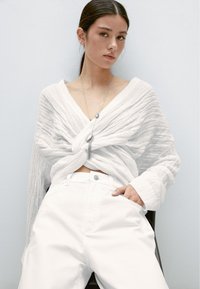 Vit texturerad omlottopp med djup V-ringning, markerad i midjan, kombinerad med vita byxor med hög midja. Enkel silversmycken framhäver outfiten.