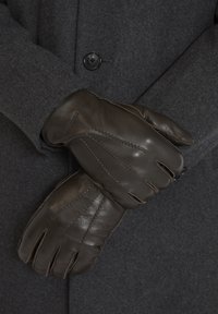 Gants en cuir marron avec des accents cousus, portés avec un manteau gris foncé. Les gants sont ajustés et ont une texture lisse.