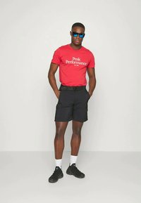 Červené bavlnené tričko s textom "Peak Performance", čierne cargo šortky, čierne tenisky a modré slnečné okuliare. Jednoduchý dizajn s voľným strihom.