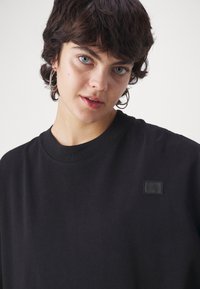 Camiseta de algodón negra con cuello redondo y mangas cortas, que presenta un pequeño parche de logotipo en relieve en la parte izquierda del pecho.