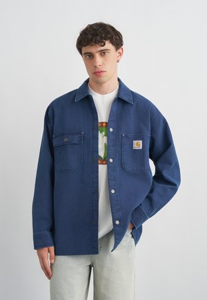 Mladý muž s krátkymi kučeravými vlasmi, oblečený v modrej kordovej bunde cez biele tričko s grafikou a svetlé rifle, stojaci pred jednoduchým pozadím.