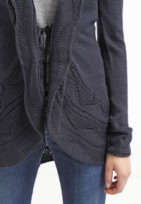 Námořnický pletený cardigan s propracovanými vlnkovými vzory, otevřeným předním designem a dlouhými rukávy. Textura je měkká, doplněná šedým tričkem pod ním.
