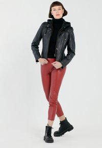 Schwarze Lederjacke mit Kapuze, mit Reißverschlüssen ausgestattete Taschen, figurbetonte rote Kunstlederhose und schwarze Schnürstiefel. Einfache Kulisse.