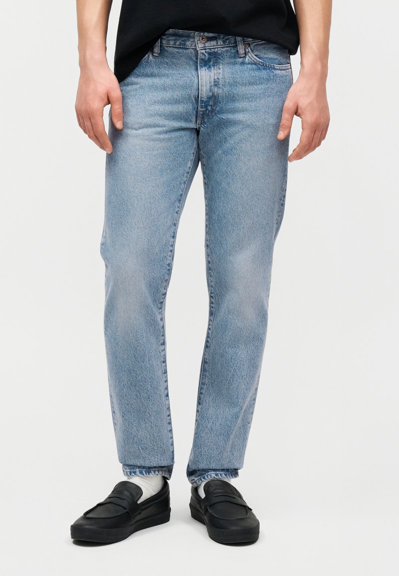 Homme portant un jean bleu clair à jambes droites, des chaussettes blanches, des mocassins noirs et une chemise noire rentrée, debout devant un fond uni.