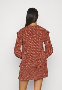 Rotes Oberteil mit gemusterten Details, Rüschenschultern und einem Peplum-Saum, kombiniert mit einem passenden, fransigen Rock. Glatte Textur, figurbetonte Silhouette.