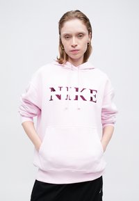 Rozā Nike kapuce ar frontālu kangaroo kabatu, ar stiepjamu kapuci un lielu, teksturētu logo tumši rozā krāsā. Mīksta auduma, izslābta piegriezuma.