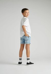 T-shirt en coton blanc, short en denim bleu clair avec des ourlets retroussés, baskets noires, et chaussettes blanches avec des rayures noires, se tenant devant un fond gris.