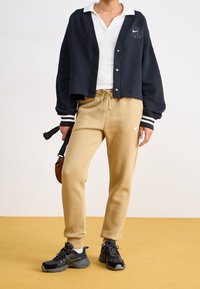 Avklippt svart kofta med vita ränder, vit pikétröja, beige mjukisbyxor och svarta sneakers med grå detaljer. Avslappnad sportig outfit.