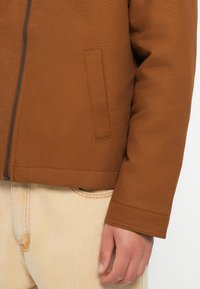 Brown jacket with a zip closure, featuring a side pocket and textured fabric. Paired with light-colored denim at the waist.
