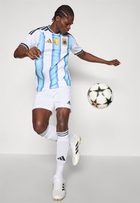 Balts futbola krekls ar zilām vertikālām svītrām, kurā ir Argentīnas emblēma. Apvienots ar baltām šortiem un melnu akcentētu apavām.