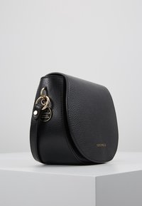 Borsa a tracolla in pelle nera a granuli con ferramenta dorata, patta arrotondata e nome del marchio "Coccinelle" impresso sul davanti.
