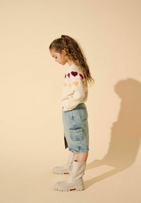 Pull en maille avec des motifs de cœurs multicolores, jupe en denim et bottes grises arrivant au genou. Le fond clair met en valeur les détails de la tenue.