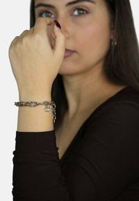 Zilveren armband met ovale schakels, een kleine bedel en een ketting sluiting, gedragen om een blote pols tegen een effen achtergrond.