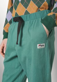 Grønne bomullsbukser med elastisk midje, svart belte, to sidelommer og et FILA-logo-lapp for merking.