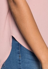 Ett arm vilar bredvid kroppen iförd en strukturerad ljusrosa topp över blå denimjeans.